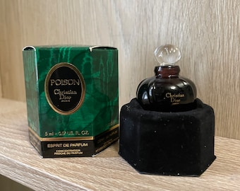 May include: A small, dark brown glass bottle of Poison perfume by Christian Dior. The bottle is in a black velvet box. The box is on a wooden surface. The bottle has a clear glass stopper. The box has a hexagonal shape. The bottle is 5 ml or 0.17 US fl oz. The perfume is an Esprit de Parfum, a concentration close to the original perfume.