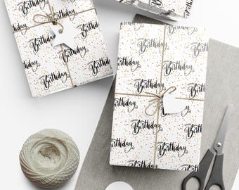 May include: White gift wrap with a repeating pattern of black script that says "Happy Birthday" and gold confetti. The gift wrap is wrapped around a box with a brown twine bow and a white gift tag.