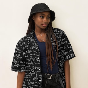 May include: A black and white short-sleeved shirt with a pattern of mathematical equations. The shirt is worn by a person with long brown braids and a black bucket hat.