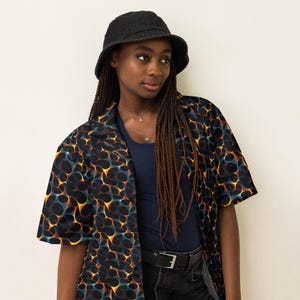 May include: A person wearing a black bucket hat and a short-sleeved shirt with a black, orange, and blue abstract pattern. The shirt has a collar and is open at the front. The person is also wearing a dark blue tank top and black jeans.