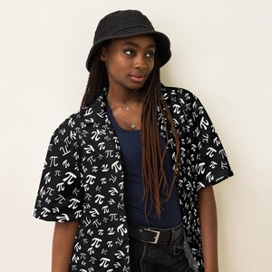 May include: A black and white short-sleeved shirt with a repeating pattern of the mathematical symbol pi. The shirt is worn by a person with long brown braids and a black bucket hat.