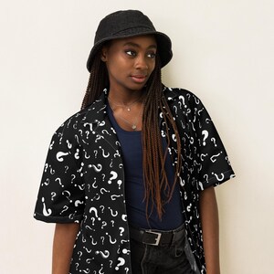 May include: A black and white short-sleeved button-up shirt with a question mark pattern. The shirt is worn by a person wearing a black bucket hat and black jeans.