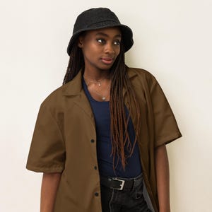 May include: A person wearing a brown short-sleeved shirt with a black button closure, a black bucket hat, and dark blue jeans. The person has long brown braids.