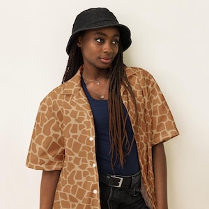 May include: A person wearing a brown and tan short-sleeved button-up shirt with a giraffe print. The shirt has a collar and a button closure. The person is also wearing a black bucket hat and a dark blue tank top.