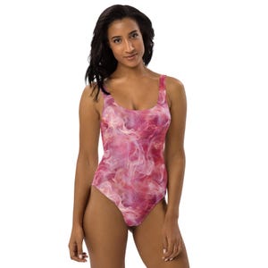 May include: A one-piece swimsuit with a pink and white abstract smoke design. The swimsuit has a scoop neckline and a low back. The fabric has a soft, flowing appearance. The model is wearing the swimsuit.