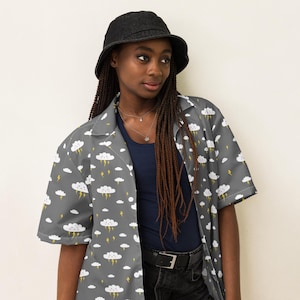 May include: A gray short-sleeved button-up shirt with a white cloud and yellow lightning bolt pattern. The shirt has a collar and a relaxed fit. The model is wearing a black bucket hat and black jeans.