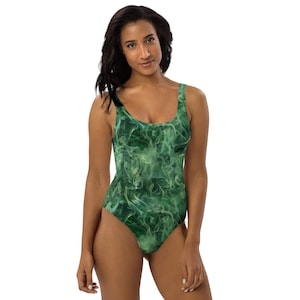 May include: One-piece swimsuit with a green and white smoke-like pattern. The swimsuit has a scoop neckline and a high-cut leg. The fabric has a smooth, shiny appearance. The overall design is abstract and artistic.