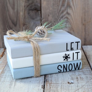 May include: Three stacked books with a rustic, farmhouse style. The top book is gray and says "LET" in black lettering. The middle book is white and says "IT" in black lettering with a snowflake symbol. The bottom book is light blue and says "SNOW" in black lettering. The books are tied together with twine and a small pine sprig.