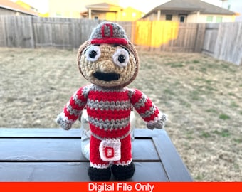 May include: A handmade crochet doll in the shape of a mascot, wearing a red and gray striped outfit and a matching hat. The doll has large, expressive eyes and a black mustache. The doll is standing on a wooden surface. The text "Digital File Only" is at the bottom.