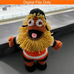 May include: A crocheted plush figure featuring a black, orange, and white color scheme with a prominent yellow yarn mane, large white eyes with dark pupils, a black face, and a small pink tongue. The text Digital File Only is at the top.