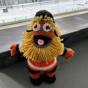 Op de afbeelding: Een gehaakt pluche speelgoed met een zwart, oranje en wit hockeytrui, een gele baard en een roze tong. Het speelgoed zit op een zwart oppervlak.