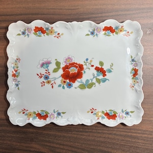 May include: A rectangular white ceramic serving tray with a scalloped edge. The tray features a central floral arrangement of red, pink, blue, and yellow flowers with green leaves, complemented by smaller matching floral clusters at each corner.