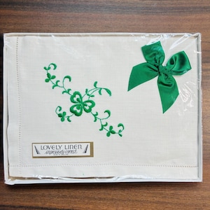 May include: A rectangular off-white linen cloth packaged in clear plastic, featuring green embroidered shamrock motifs and a decorative green satin ribbon bow. A rectangular label reads LOVELY LINEN impressively correct MADE IN IRELAND.