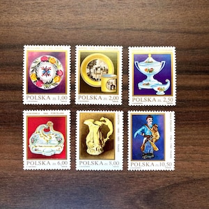 May include: A collection of six Polish postage stamps featuring images of porcelain and pottery. The stamps display colorful illustrations of plates, pitchers, and figures, with text in Polish. The stamps are arranged on a wooden surface.