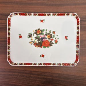 May include: A rectangular white ceramic serving tray featuring a central floral arrangement of red, yellow, and black flowers with green leaves. The border is decorated with a repeating pattern of red, blue, and gold geometric and floral motifs.