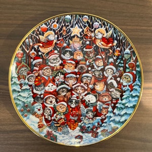 May include: A Christmas-themed ceramic plate with a gold rim, featuring a festive scene of cats dressed in Santa hats and winter clothing, gathered around a Christmas tree with ornaments and presents. The plate has a white background with a blue and white snowy landscape.
