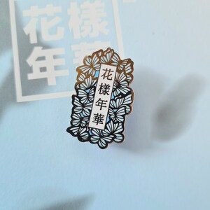 以下が含まれることがあります： 青と白の花柄の金色の金属製のピン。ピンには、黒い文字で「花樣年華」と書かれた白い長方形があります。