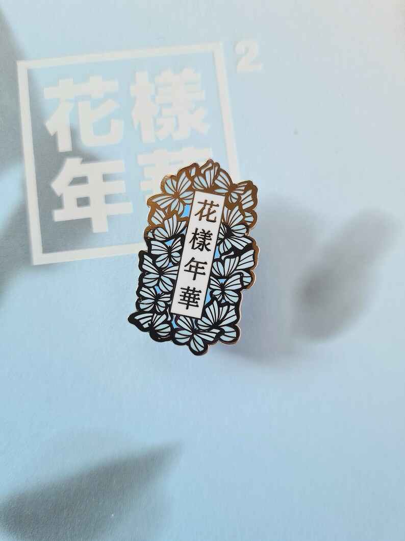 以下が含まれることがあります： 青と白の花柄の金色の金属製のピン。ピンには、黒い文字で「花樣年華」と書かれた白い長方形があります。