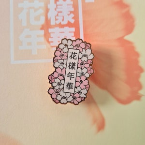 以下が含まれることがあります： 白い背景にピンクの桜の花が咲いた金色のエナメルピン。ピンには黒い文字で「花樣年華」と書かれている。