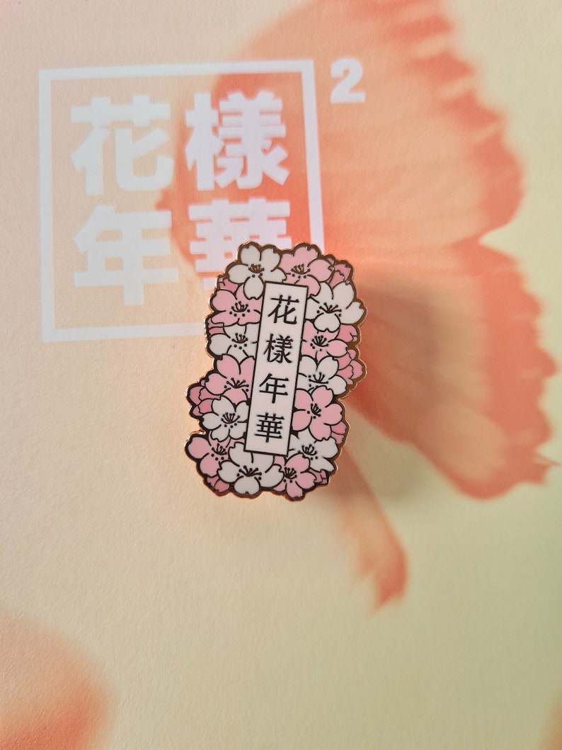 以下が含まれることがあります： 白い背景にピンクの桜の花が咲いた金色のエナメルピン。ピンには黒い文字で「花樣年華」と書かれている。