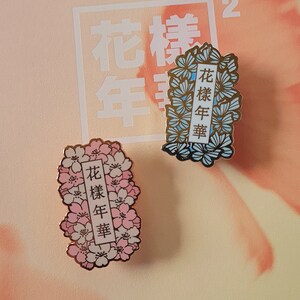 以下が含まれることがあります： 花柄のデザインが施されたエナメル製のピンが2つ。ピンには「花樣年華」という文字が金色の文字で書かれている。1つのピンはピンクと白の花柄、もう1つのピンは青と白の花柄である。