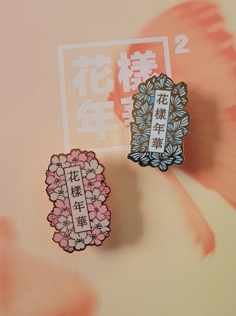 以下が含まれることがあります： 花柄のデザインが施されたエナメル製のピンが2つ。ピンには「花樣年華」という文字が金色の文字で書かれている。1つのピンはピンクと白の花柄、もう1つのピンは青と白の花柄である。