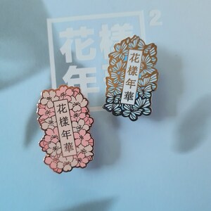以下が含まれることがあります： 花柄のデザインが施されたエナメル製のピンが2つ。ピンは金色で、青とピンクの花が描かれている。ピンに書かれている文字は中国語で「花樣年華」である。