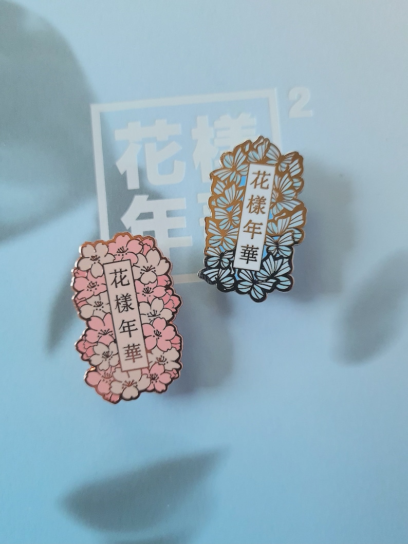 以下が含まれることがあります： 花柄のデザインが施されたエナメル製のピンが2つ。ピンは金色で、青とピンクの花が描かれている。ピンに書かれている文字は中国語で「花樣年華」である。