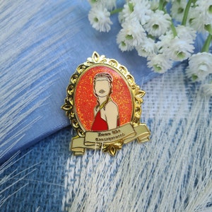 May include: Gold-toned enamel pin featuring a woman with red hair in a red dress, set against a red glitter background. The pin has a banner that reads "Down the Consequences".