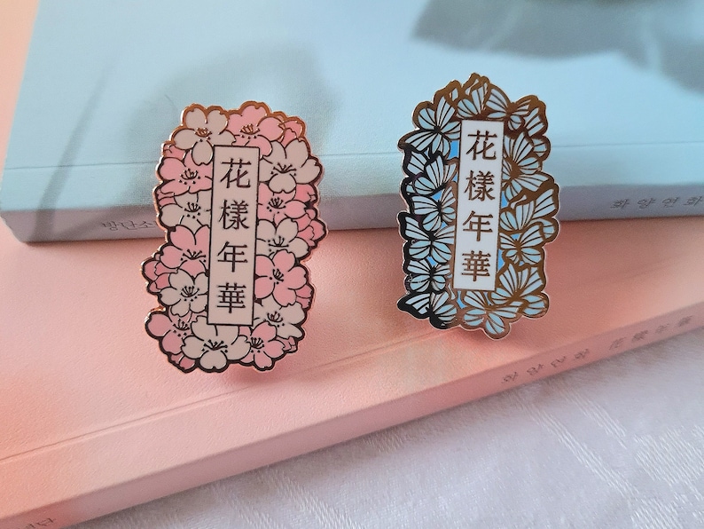 以下が含まれることがあります： 花柄のデザインが施されたエナメル製のピンが2つ。ピンクのピンは桜の花と金色の文字で「花樣年華」と書かれている。青いピンは蝶と金色の文字で同じ文字が書かれている。