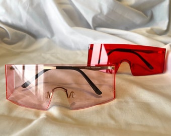 May include: Two pairs of rectangular sunglasses with a flat top and curved bottom. The sunglasses are made of a clear plastic material. One pair is red and the other is pink. The sunglasses are lying on a white fabric surface.