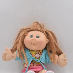 May include: A vintage Cabbage Patch Kids doll with red hair and green eyes. The doll is wearing a blue cardigan, a floral top, and a pink skirt. The doll's shoes are white with colorful accents. The doll's arms are outstretched.