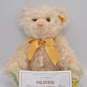 May include: A light beige mohair teddy bear with a yellow satin ribbon around its neck. The bear has dark eyes, a stitched nose, and a yellow tag in its ear. One foot pad features green musical notes, and the other features yellow daffodils. A certificate of authenticity for Oliver The Musical Spring Bear is placed in front.