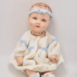 May include: A vintage doll with a painted face, wearing a white knitted outfit with blue accents. The doll has a matching blue headband and blue knitted booties. The doll is sitting down.