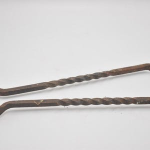 Pode incluir: Um par de tenazes de ferro enferrujado com um cabo em forma de laço e dois braços longos com um design de metal torcido. As extremidades dos braços são moldadas com pequenos dentes e o objeto tem um acabamento de metal oxidado castanho.
