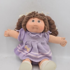 May include: A vintage Cabbage Patch Kid doll with brown yarn hair in pigtails. The doll is dressed in a lavender knit dress with a duck applique and white socks. The doll is sitting with arms outstretched.