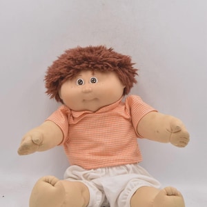 May include: A vintage doll with brown hair, freckles, and round eyes. The doll wears an orange and white checkered shirt and white shorts. The doll's arms and legs are outstretched, and it is sitting down.