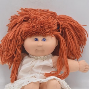 May include: A vintage doll with orange yarn hair and blue eyes. The doll wears a white dress with a lace trim. The doll's arms and legs are made of a soft material.