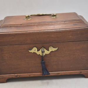 Puede incluir: Una caja de madera con asa y herrajes decorativos de latón. La caja es rectangular con tapa abisagrada y acabado marrón oscuro. Una borla azul oscuro cuelga del ojo de la cerradura.