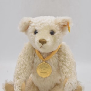 Puede incluir: Un oso de peluche color crema con una medalla dorada alrededor del cuello. El oso tiene ojos oscuros y una nariz marrón claro. El oso está sentado con las piernas extendidas. La medalla tiene texto.