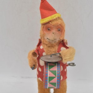 May include: Vintage wind-up toy monkey wearing a red polka-dot vest and a matching hat with yellow trim. The monkey is holding a drum and drumsticks, a classic collectible toy.