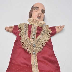 Könnte beinhalten: Eine Vintage-Puppe, die William Shakespeare darstellt. Die Puppe hat ein bemaltes Gesicht, eine Glatze und braunes Haar. Sie trägt ein rotes Kleid mit dekorativem Kragen und Besatz. Die Arme der Puppe sind ausgestreckt.