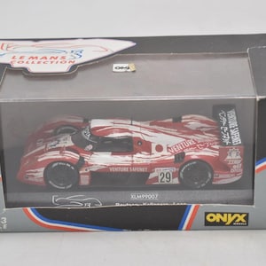 Op de afbeelding: Een 1:43 schaalmodel raceauto in een doorzichtige display case. De auto is rood en wit met sponsorlogo's en het nummer 29. De doos heeft het "Le Mans Collection" logo en het merk Onyx Models.
