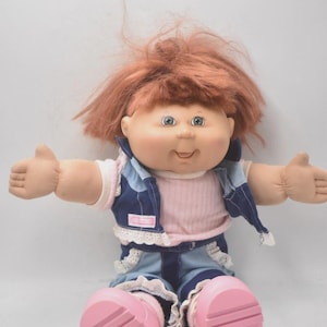 Op de afbeelding: Een vintage pop met rood haar, blauwe ogen en een glimlachende uitdrukking. De pop draagt een roze shirt, een spijkerbroekvest en -short met witte kanten rand en roze schoenen. De armen van de pop zijn uitgestrekt.