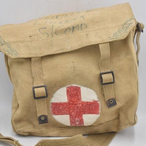 May include: Tan canvas medical bag with a red cross emblem. The bag has a shoulder strap, two front straps with buckles, and text on the flap. The bag is likely vintage or military-inspired.