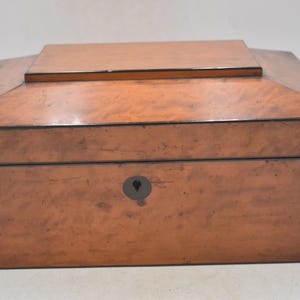 Peut inclure: Une boîte en bois de forme rectangulaire avec un couvercle à charnière. Le bois est brun clair avec des bordures marron foncé et un trou de serrure à l'avant. Le dessus est légèrement incliné, avec un petit couvercle rectangulaire.