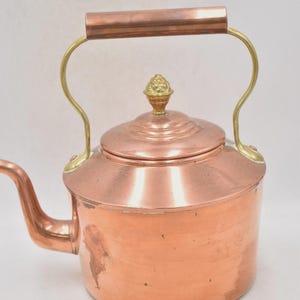 Può includere: Una teiera in rame con manico e beccuccio in ottone. La teiera ha un coperchio con un pomello decorativo in ottone. Il manico è curvo e fissato al corpo con raccordi in ottone. La teiera è progettata per l'uso sul piano cottura.