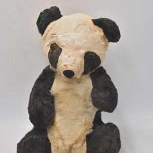 May include: A well-worn panda bear soft toy, featuring black and cream colours. The bear has black ears, eyes, and limbs, with a cream-coloured face and body. The soft toy is in a sitting position and shows signs of age.