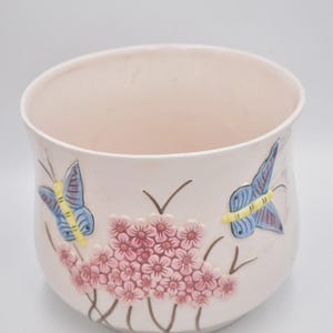 May include: A pale pink ceramic plant pot with a wide opening. It features raised relief designs of pink flowers and blue and yellow butterflies. Suitable for indoor or outdoor use, it measures approximately 20 cm in diameter.