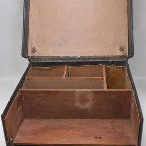 Pode incluir: Uma mala de couro preta vintage aberta, com interior castanho claro. A mala tem vários compartimentos e prateleiras de madeira, sugerindo que foi projetada para armazenamento ou organização. A mala está aberta, revelando sua estrutura e design interior.